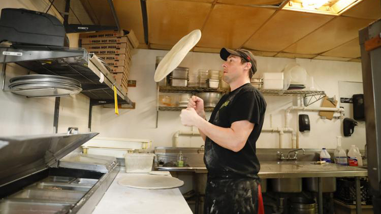 Dennis Laubner hand-tosses a pizza, facing left towards a counter in the kitchen of Monona Pizza and Subs